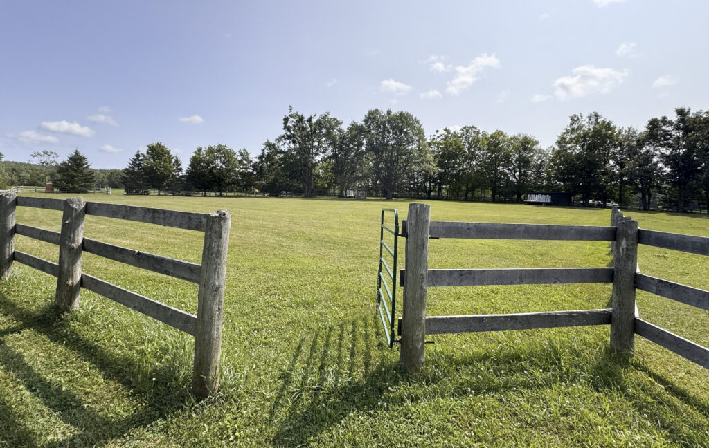 Open pasture field at Glen Oro Farm with a wooden split-rail fence and gate, showing jump obstacles in the distance