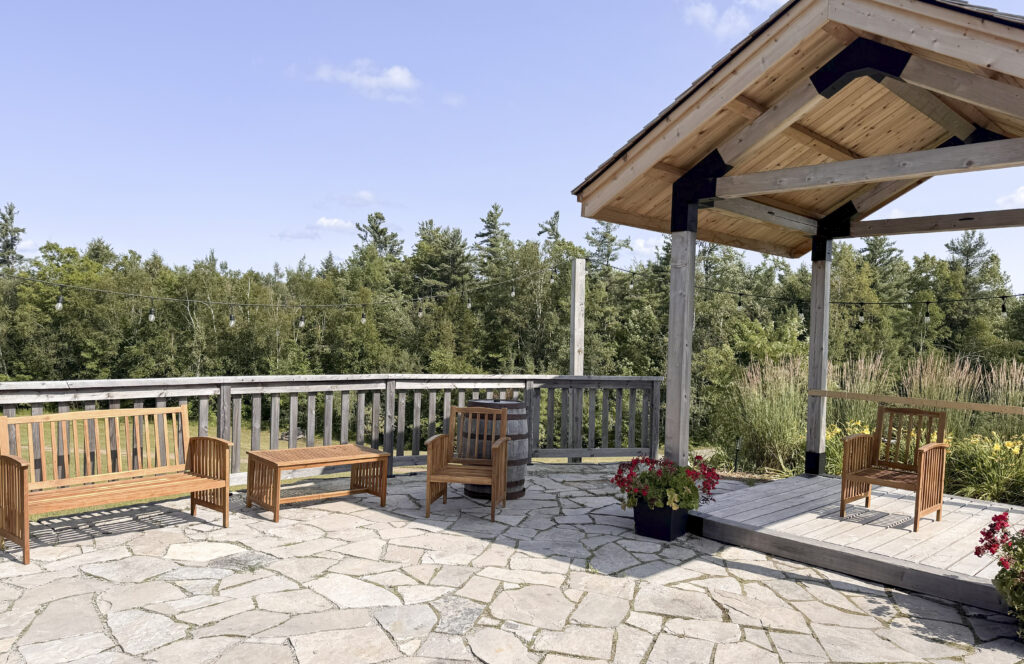 Covered wooden pergola with lounge seating on a stone patio at Glen Oro Farm, overlooking the tree line