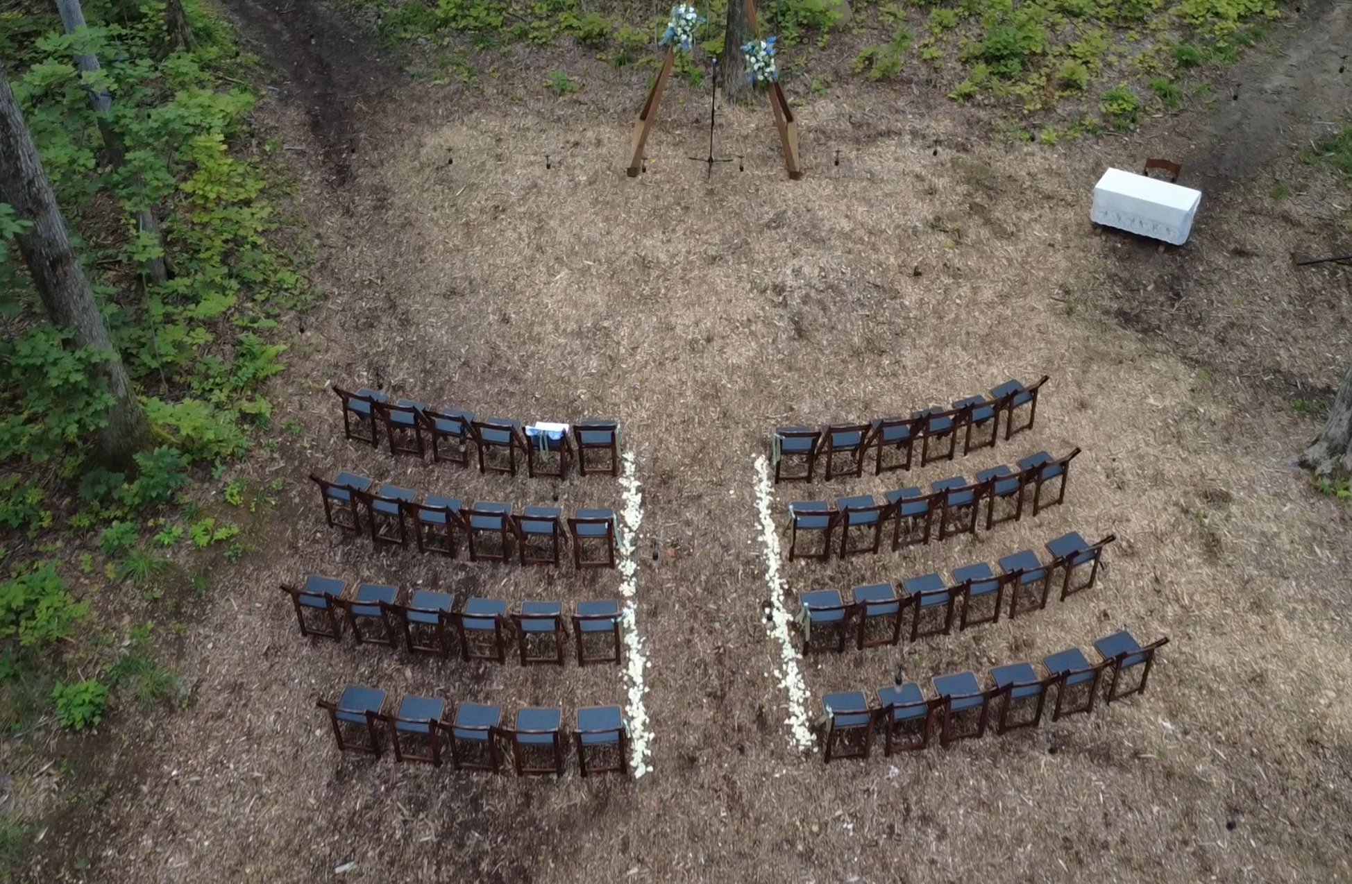 Aerial view of The Sanctuary forest ceremony space at Glen Oro Farm, showing rows of chairs split by a petal-lined aisle leading to a floral arbor among the trees.